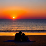 Sunset on the beach. A couple lays on a colorful towel on the sand. The vantage point is from the couple looking out, so only their legs on the towel are showing with all of the scenery