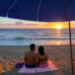 Sunset on the beach. A couple lays on a colorful towel on the sand. The vantage point is from the eyes of the couple looking out, so only their legs on the towel are showing with all of the scenery