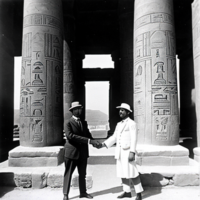 1920s black and white photograph of two explorers, shaking hands in front of an Egyptian temple with hieroglyphics on the walls